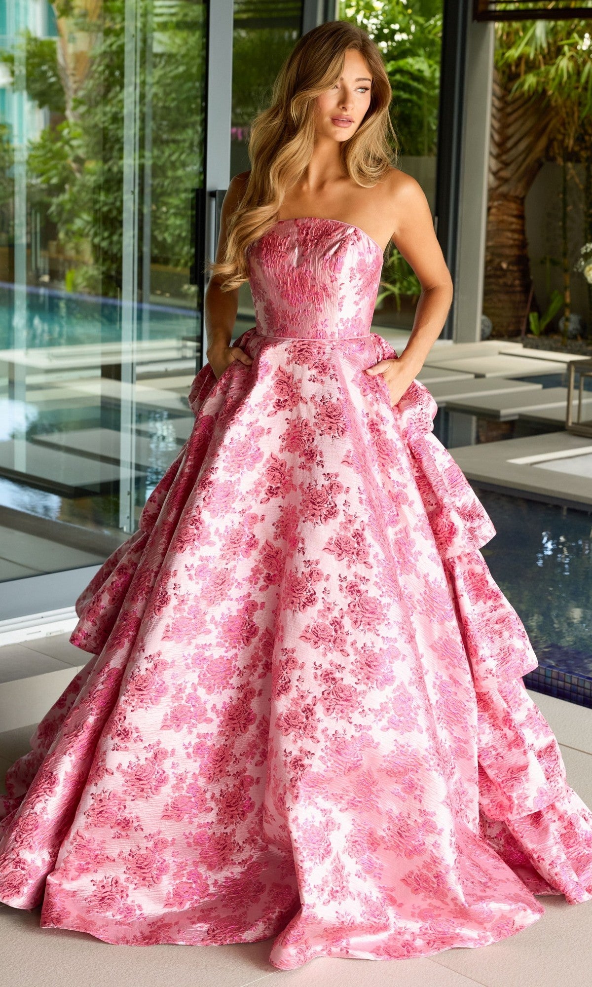 Indoors by a pool, a woman wears the Ruffled Strapless Prom Gown: Ava Presley 26412, an A-line prom dress with pink floral print, full skirt with pockets, and elegant back ruffles.